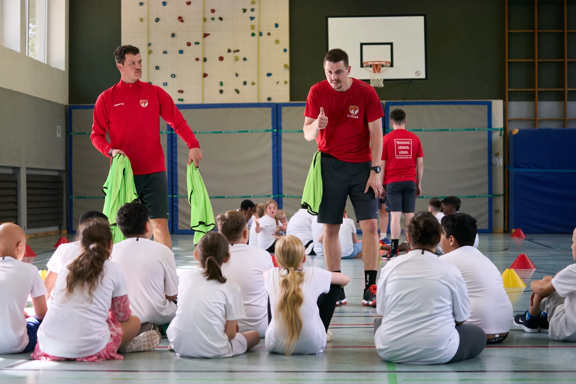 Zwei Trainer in roten Trikots erklären einer Gruppe von Kindern in weißen Trikots, die im Kreis auf dem Hallenboden sitzen, die nächste Übung.